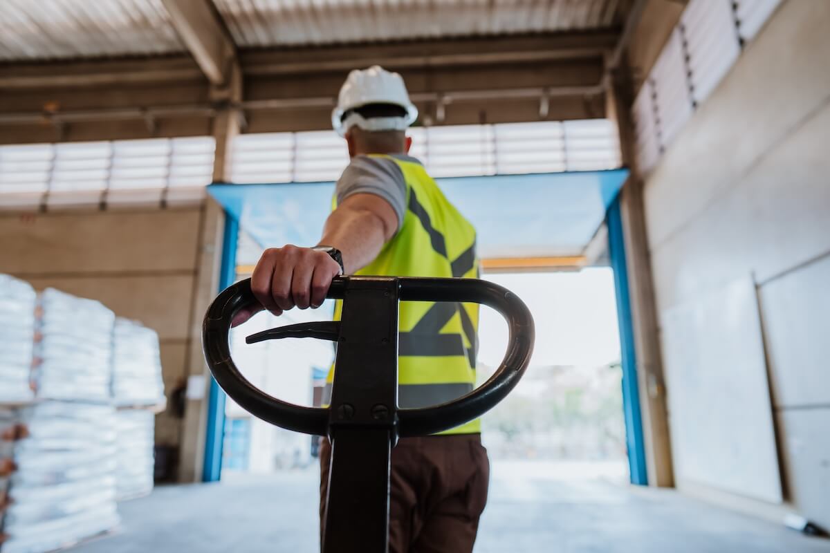 Temporary industrial staff worker wearing hard hat and vest pulling dolly.